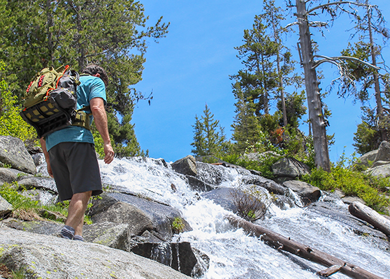 Hiker wearing a green backpack crossing a waterfall